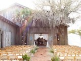 a simple and cool outdoor barn space with white curtains, monograms on the doors, potted plants that line up the wedding aisle