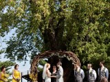 a rustic outdoor wedding ceremony space with a vine wedding arch with greenery and blooms and blooms in jars lining up the wedding aisle