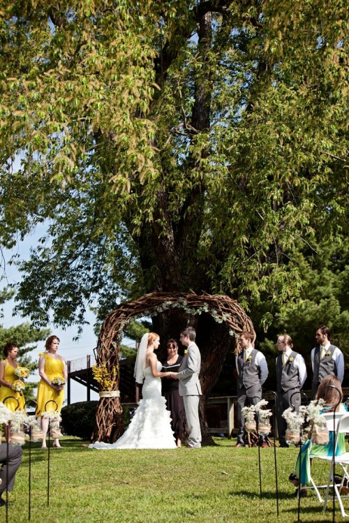a rustic outdoor wedding ceremony space with a vine wedding arch with greenery and blooms and blooms in jars lining up the wedding aisle