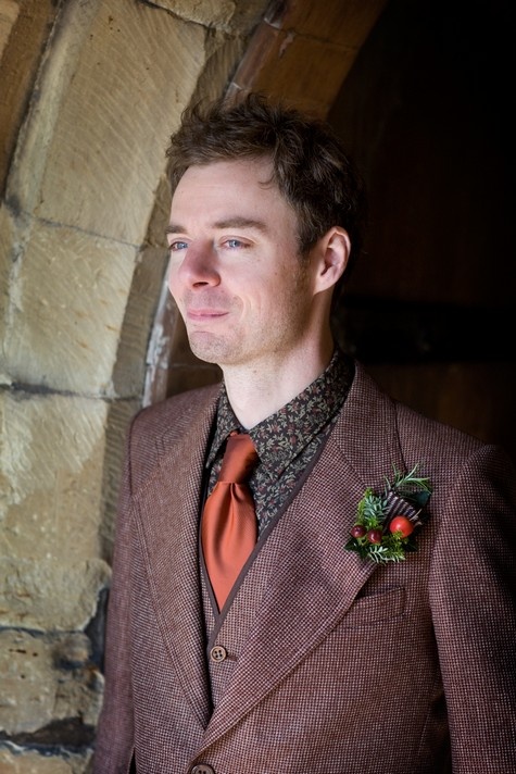 a burgundy tweed three piece suit, a moody floral shirt, a rust tie and a berry boutonniere