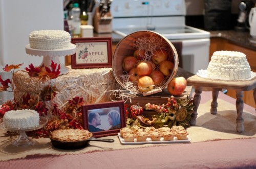 a fall bridal shower dessert table with cute cakes, tarts, apples and cupcakes plus hay for a rustic feel