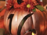 a metallic rust-colored faux pumpkin with a cutout number, a candle inside and bright faux blooms on top