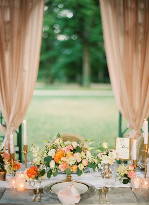 a romantic and elegant wedding tablescape with a peach, orange and white floral centerpiece, candles, blush candleholders and a blush napkin