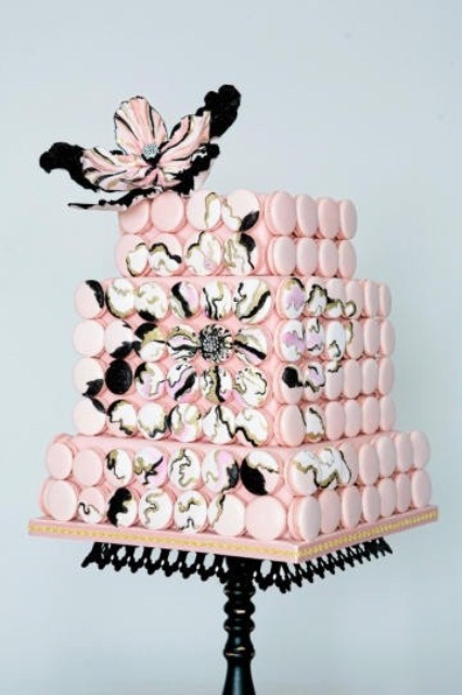 a square pink wedding cake covered with pink and painted macarons that show off blooms, with pink, white and black sugar blooms on top