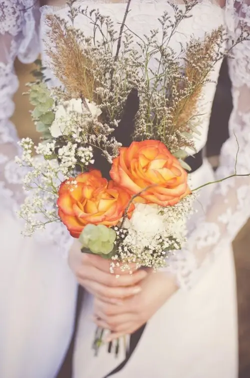 a fall boho wedding bouquet with orange blooms, baby's breath, dried elements and grasses plus some greenery