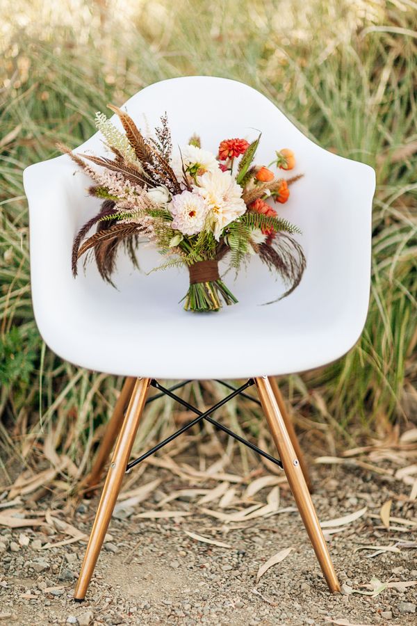 A bright fall boho wedding bouquet of neutral and orange blooms, greenery, feathers, dried touches and with a brown wrap