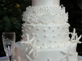 a white shimmering wedding cake decorated with edible pearls, corals, starfish and pearls and starfish on top is very elegant