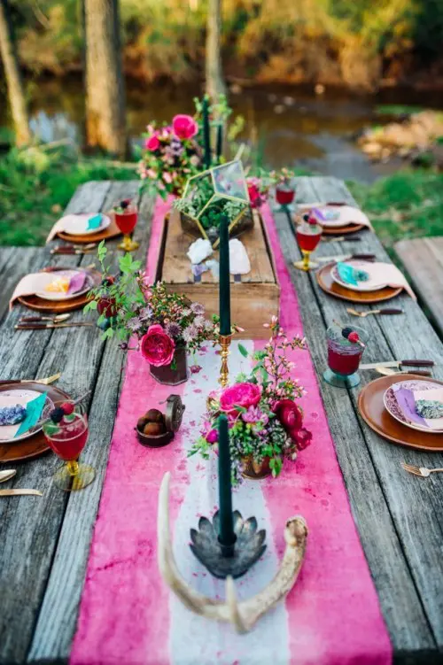 a bold boho woodland wedding tablescape with a colorful runner, antlers, dark candles and old blooms plus gold cards and wood print chargers