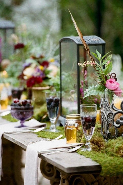 a decadent woodland boho wedding table with a moss runner, candle lanterns, bold blooms and fruit and berries served