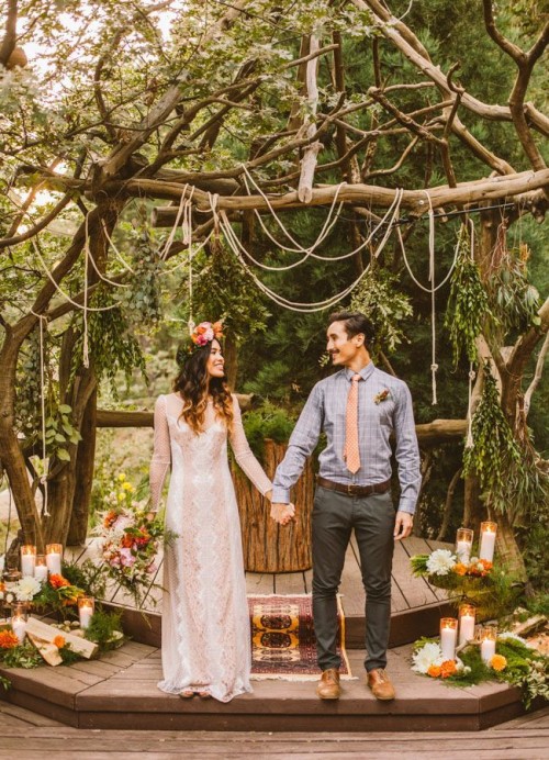 a boho woodland wedding altar done with lots of candles, greenery, bold blooms, ropes hanging over it and some boho rugs