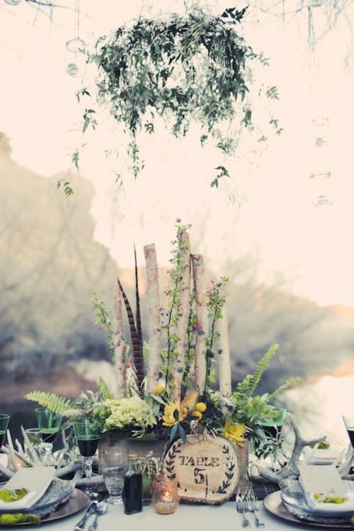a boho woodland wedding tablescape with a crate with branches, greenery, blooms and feathers, a greenery wreath over the table, antlers, green glasses and a wood burnt table number