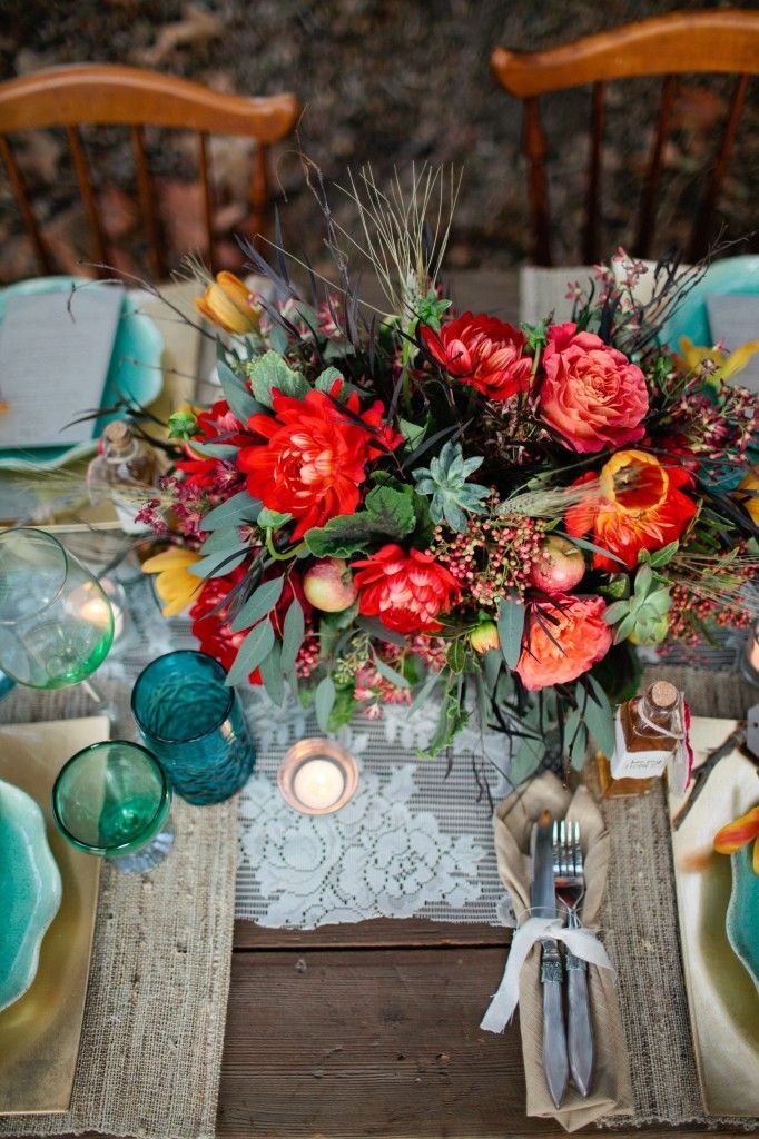 A bright boho chic wedding tablescape with a lace runner and burlap placemats, a bold floral centerpiece with greenery, candles and blue and green glasses