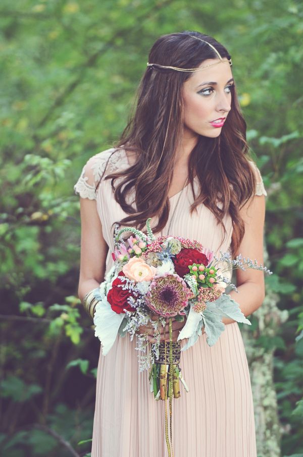 A boho woodland bride wearing a blush pleated wedding dress with lace cap sleeves, a boho chain on the head and carrying a very bold floral bouquet