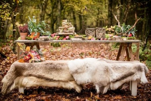 a boho woodland wedding tablescape with a fur covered bench, potted greenery, antlers, woven candle lanterns, fruits and leaves is a lovely and chic idea