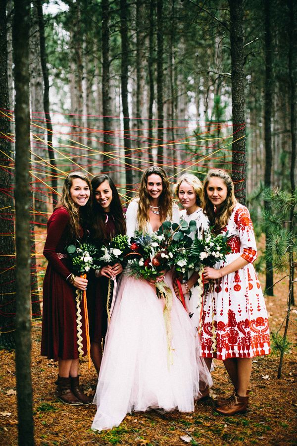 A boho woodland wedding backdrop of colorful yarn put on living trees and boho bridesmaids wearing mismatching burgundy and white dresses