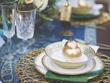 a refined woodland wedding place setting with a gold woven placemat, green and blue linens, gilded pears and bold blooms and greenery