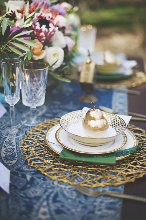 a refined woodland wedding place setting with a gold woven placemat, green and blue linens, gilded pears and bold blooms and greenery