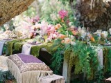 a boho woodland wedding tablescape with a moss tablecloth, lavender linens, bright blooms and lots of ferns plus purple boho pillows