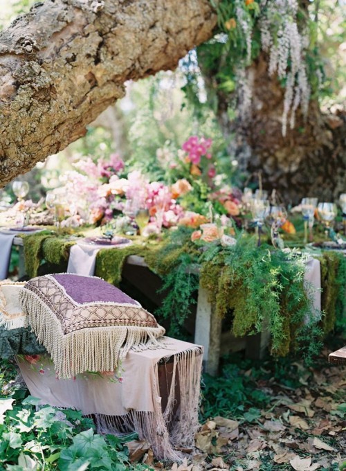 a boho woodland wedding tablescape with a moss tablecloth, lavender linens, bright blooms and lots of ferns plus purple boho pillows