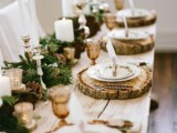 a cozy rustic woodland wedding tablescape with wood slice placemats, an evergreen runner with pinecones, gilded tuches and amber glasses