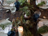 a simple woodland tablescape with an uncovered table, a textural greenery table runner, blue glasses and ferns on each place setting