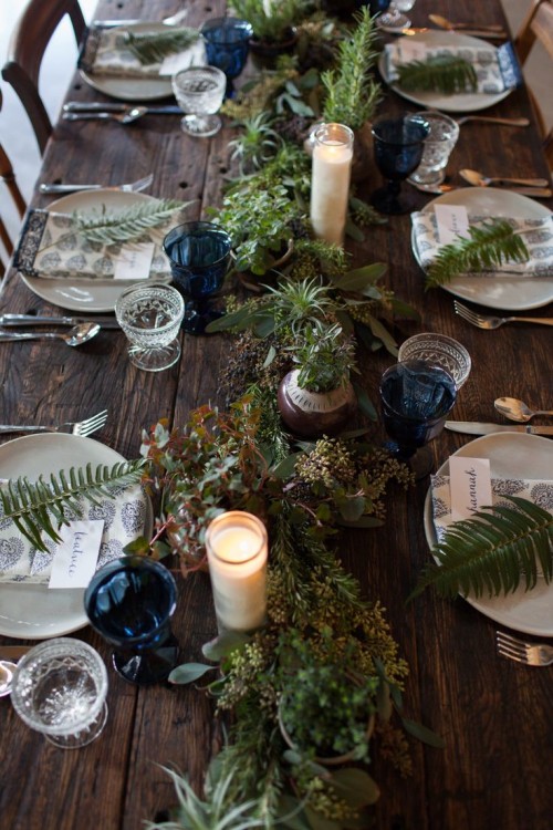 a simple woodland tablescape with an uncovered table, a textural greenery table runner, blue glasses and ferns on each place setting