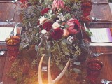 a boho woodland tablescape with an uncovered table, a greenery runner and blooms, candles and antlers