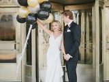 the groom wearing black, the bride wearing white and gold and black balloons