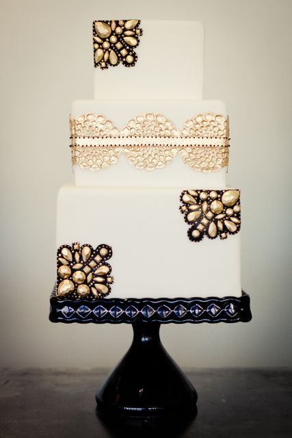 a white wedding cake decorated with gold edible lace and gold rhinestones and black lace