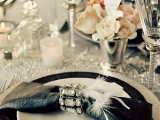 a chic black, white and silver wedding tablescape with various plates, a black napkin with a rhinestone ring, feathers and a sparkling table runner
