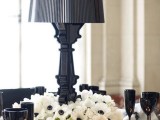 a black and white tablescape with a floral centerpiece and a black lamp, black glasses, white candles and black napkins