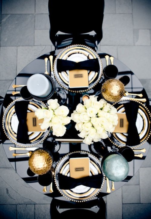 a holiday black, white and gold wedding tablescape with large gold ornaments and cutlery, neutral blooms and a large candle