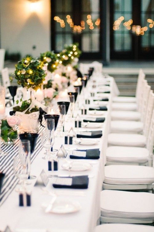 a contemporary black and white tablescape with a striped table runner, neutral blooms and greenery topiaries with lights, black glasses and napkins