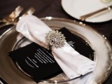 a black and white place setting with silver touches, a large brooch napkin ring and a dark tablecloth