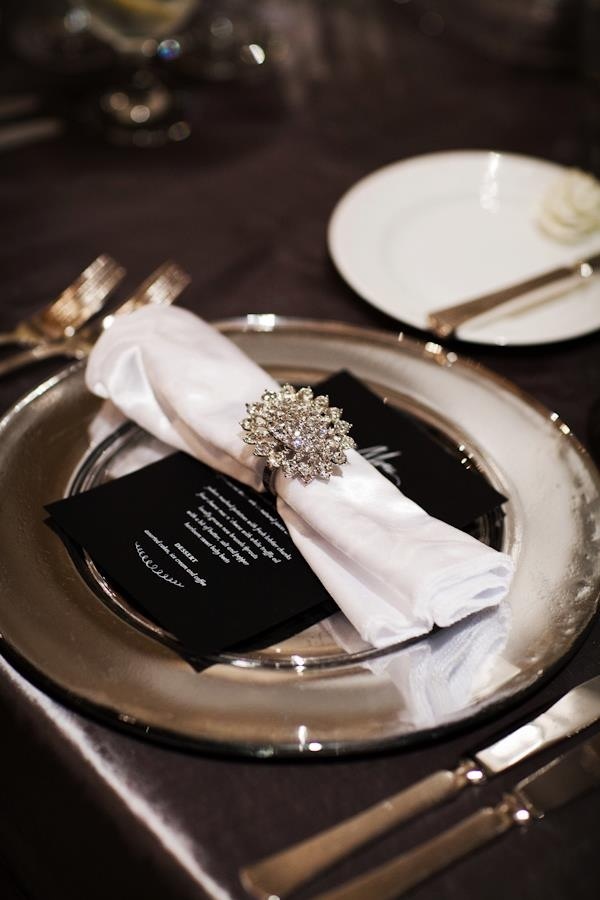A black and white place setting with silver touches, a large brooch napkin ring and a dark tablecloth