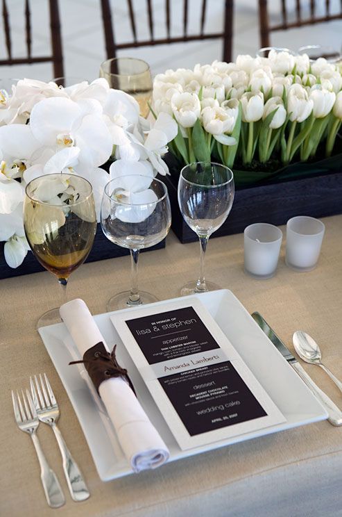 a super elegant black and white table setting with a square plate, a black and white menu, a white napkin, white orchids and tulips