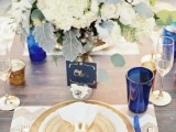 a chic place setting with a chevron placemat, a gold and white setting, blue glasses and vases and a lush centerpiece