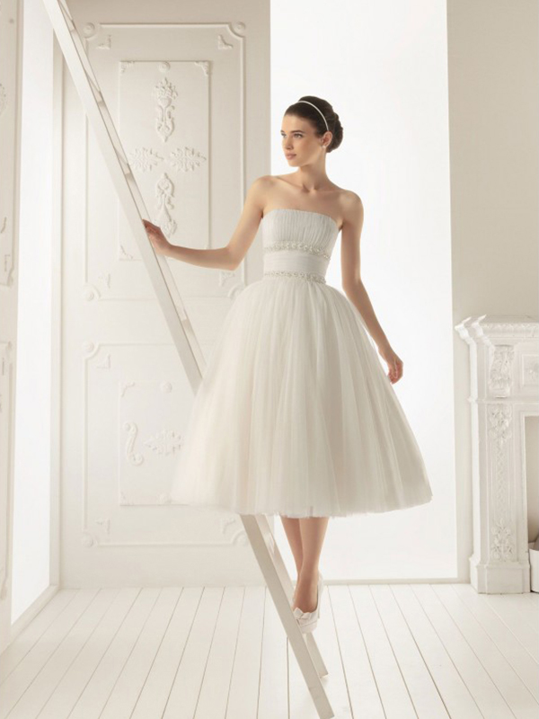 A strapless A line tea length wedding dress with a pleated bodice, a wide sash and a pleated skirt, white shoes and a headband