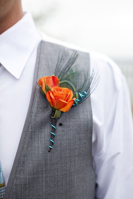 a catchy wedding boutonniere of orange roses, feathers and a wrap is a stylish touch of color to the groom's look
