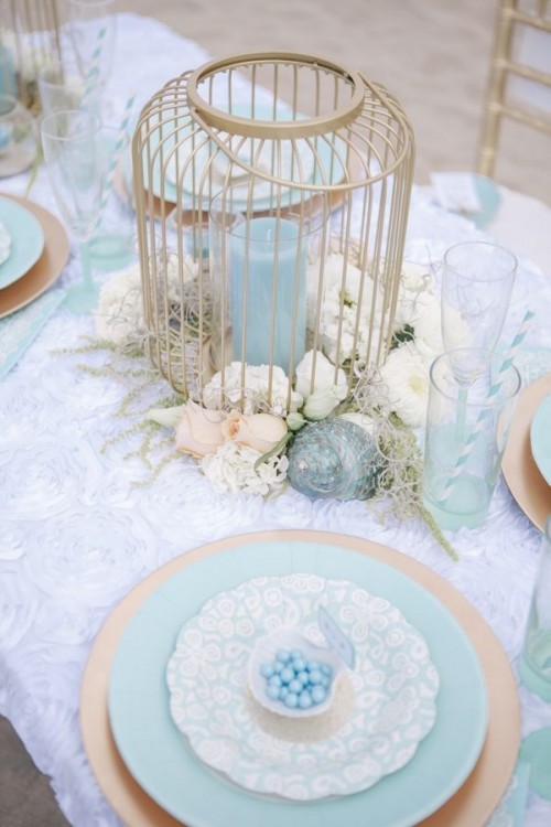 a pastel ethereal bridal shower tablescape with white and blue linens, a cage with a blue candle, white blooms, seashells and blue plates looks veyr beautiful
