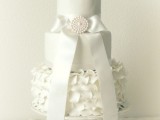 a white wedding cake with plain and a ruffle tier, with a large ribbon bow and a brooch, white sugar blooms on top