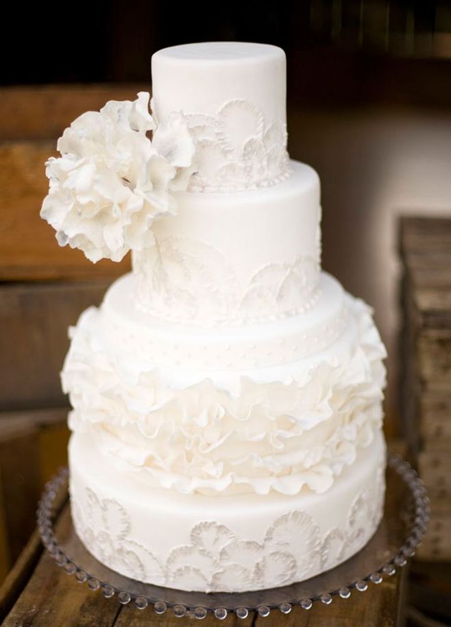 A refined white patterned wedding cake with floral and ruffles patterns and oversized white sugar blooms on top