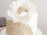 a white wedding cake with gold detailing and an oversized white sugar bloom looks very chic and statement-like