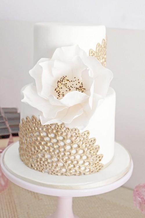 a white wedding cake with gold detailing and an oversized white sugar bloom looks very chic and statement-like