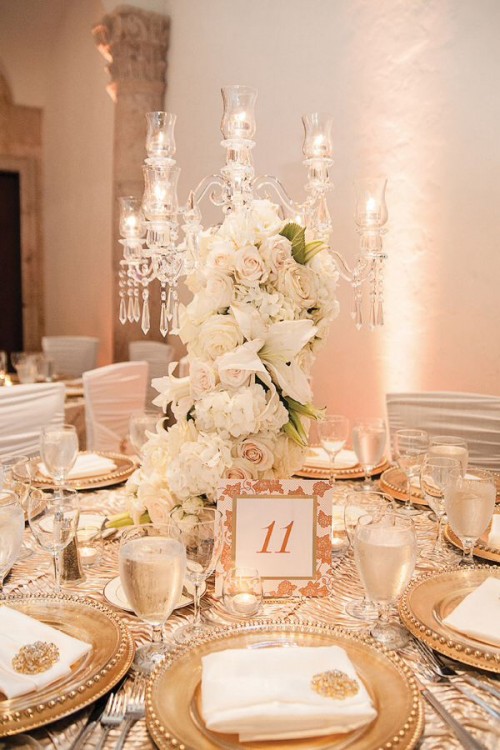 a chic wedding tablescape in gold and white, with a refined crystal centerpiece with white blooms, gold chargers and cutlery plus white and gold linens