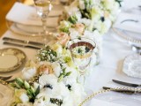 an exquisite gold and white wedding tablescape with a white bloom table runner, gold rimmed glasses and chargers and gold cutlery