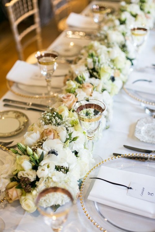 an exquisite gold and white wedding tablescape with a white bloom table runner, gold rimmed glasses and chargers and gold cutlery