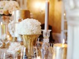 a refined gold and white wedding tablescape with gold and blush candles, glasses, an embellished tablecloth and white blooms in gold vases