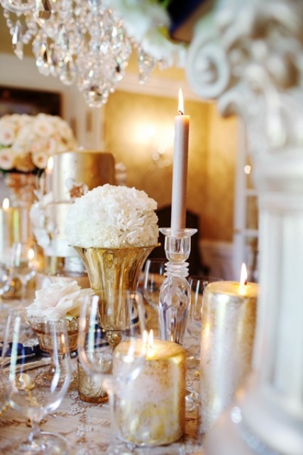 a refined gold and white wedding tablescape with gold and blush candles, glasses, an embellished tablecloth and white blooms in gold vases