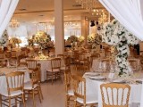 a gold and white wedding reception space with white floral arrangements, white linens and gold chargers and cutlery plus gold chairs